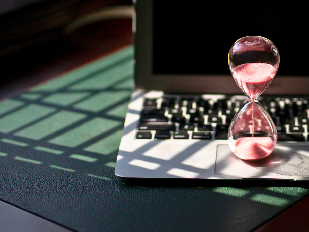 A glass sand timer resting on a laptop with a dark green shadowy background.